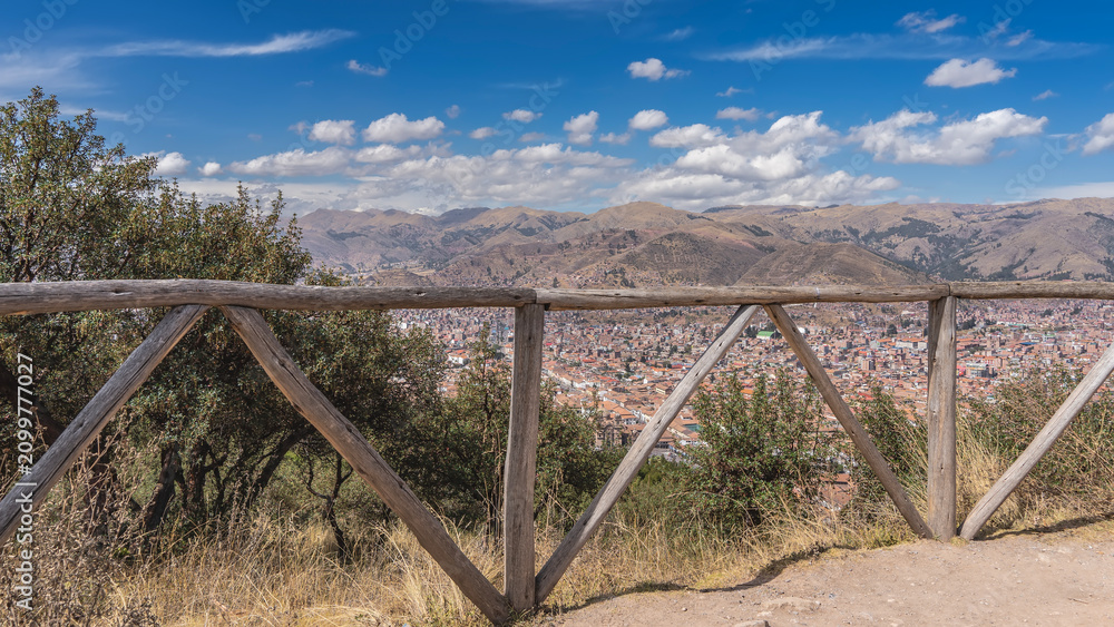 Fototapeta premium High-altitude observation deck. Behind the wooden railings, you can see the city from a height. Lots of buildings with red tiled roofs. Mountains against a blue sky and clouds. Peru. Cusco.