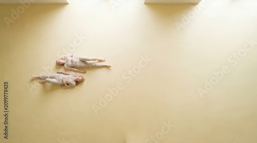 Two women lie down on pastel yellow wall viewed from above surreal empty space.