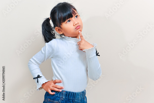 Bild auf Leinwand A cute little Asian girl with pigtails hair looking up thoughtfully while holding her chin with a hand against an isolated background