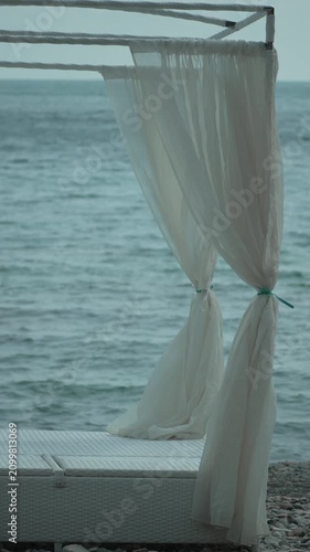 Beach canopy ocean, white bed with flowing curtains by calm sea, serene vacation spot. Vertical video.