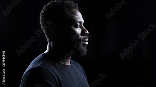 Man sitting upright in complete darkness, only faint light catching the edge of his features. Deep introspection concept