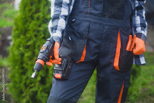 person holding power drill outdoors wearing orange gloves and black jacket for home improvement project in garden setting close-up practical and serene mood natural light clean background.