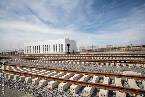 New train tracks with concrete sleepers and gravel ballast near railway building.
