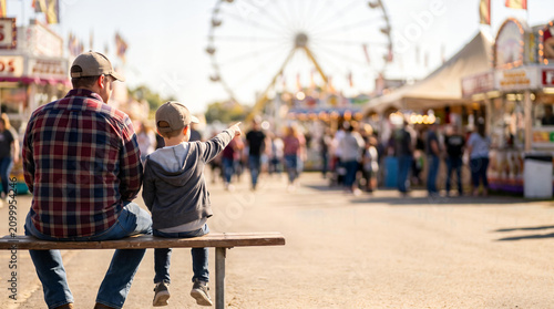 Rear view of a father and son sitting on a wooden bench pointing at a Ferris wheel at a summer county fair, featuring blurred background and ample copy space