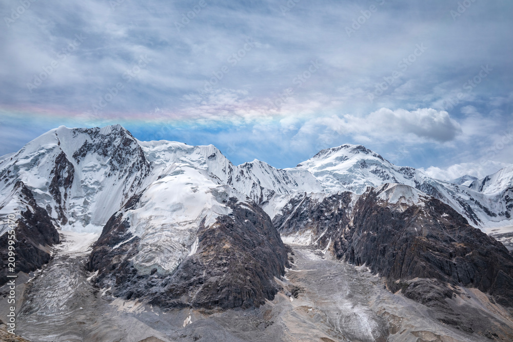 Obraz premium A stunning rainbow arches over the snow-capped peaks of the Tianshan Mountains in Xinjiang, China, creating a breathtaking natural spectacle against a dramatic sky.