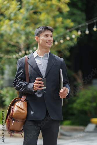 Asian businessman walking with laptop and coffee outdoors