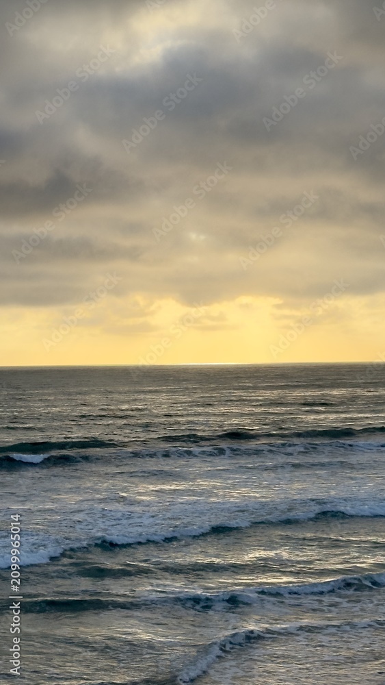 Fototapeta premium A portrait layout vertical panorama capturing layered grey storm clouds over golden light reflecting on rolling ocean swell waves in Carlsbad, California.