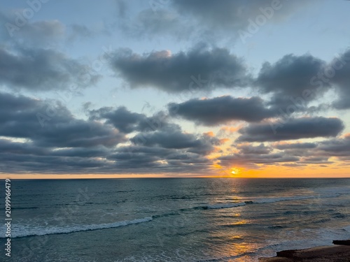 Фотография A wide seascape showcasing a vibrant orange sunset on the horizon with a gentle ocean swell rolling across the foreground at Carlsbad State Beach, California