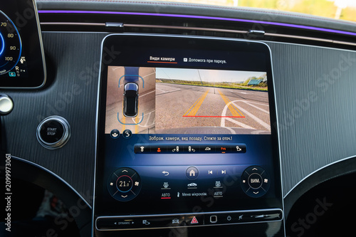 Large touchscreen infotainment display in a luxury car showing a 360-degree backup camera view and parking assist system with active guidelines, close up.