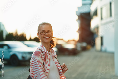 A smiling young woman in striped shirt with glasses walks along a sunny urban street  radiating confidence and calm  ready for a casual day.