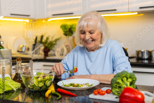 Senior old woman smiling while eating fresh vegetable salad at home kitchen for diet support. Elderly grandmother enjoying healthy safe meal choice with calm mood for wellness nutrition goal indoors.