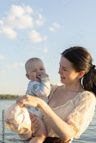 Mother holding happy baby in water during golden hour with clouds in the sky