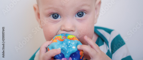 Картината върху платно Tiny child with sparkling blue eyes focuses intently while gnawing on a bright p