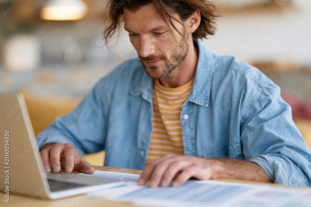 Fototapeta premium Man focused on laptop, fingers poised over keys. Casual attire, layered shirts, relaxed posture. Soft lighting, warm tones, calm indoor setting. Conveys concentration, productivity, digital engagement