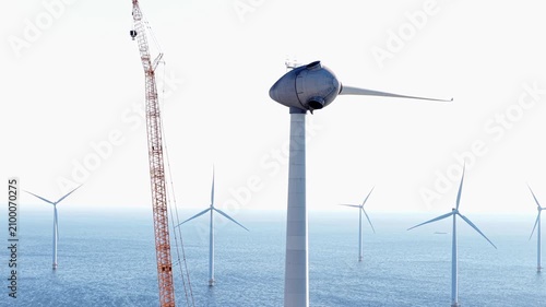 A tall orange crane stands beside an offshore wind turbine under construction in the North Sea. Several other white wind turbines rise from the water in the distance. The sky is bright and clear.