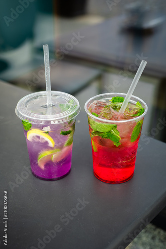 Homemade fruit lemonades in takeaway cups. Bright red strawberry lemonade and butterfly pea flower lemonade with ice, citrus and mint. Refreshing non-alcoholic summer beverages.