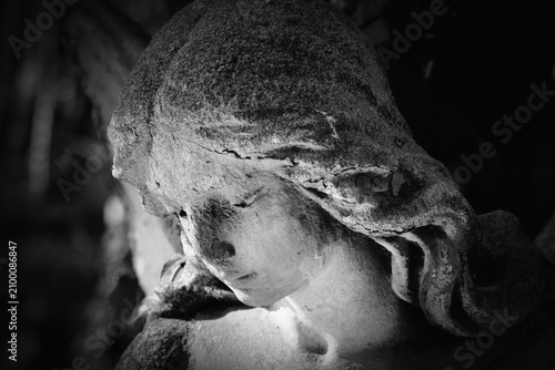 Close-up of a weathered stone angel with bowed head in black and white, expressing sorrow, remembrance, silence, and eternal peace.