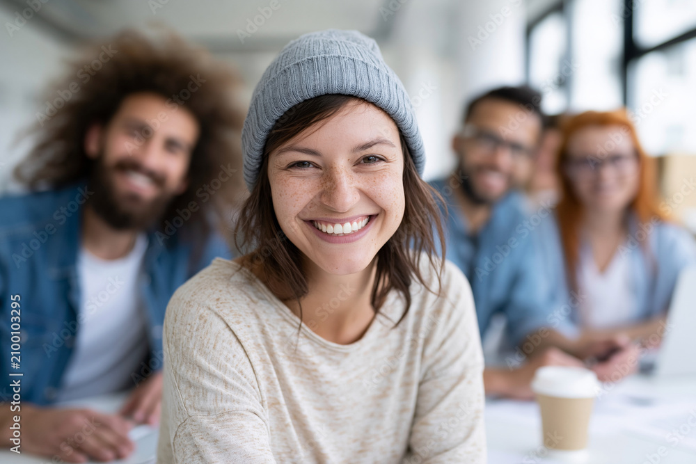 Fototapeta premium Young woman smiles warmly at camera. Colleagues sit blurred behind her in bright office. Natural light illuminates her cheerful expression. Casual attire suggests relaxed, creative workplace