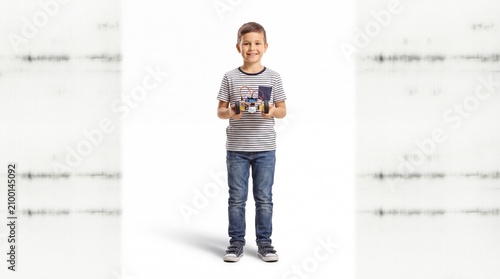 Full-length portrait of a smiling young boy in a striped t-shirt and jeans, standing on a white background and holding up a custom-built modular robotic toy car