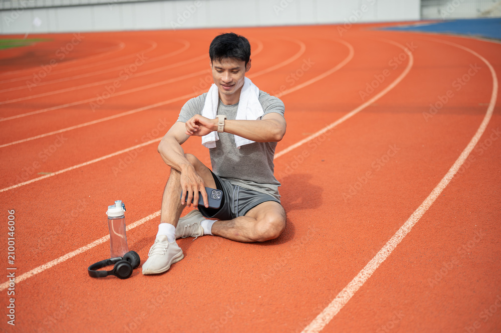 Obraz premium Asian athletic man checking time or reviewing smartwatch while recovering on a track after running.