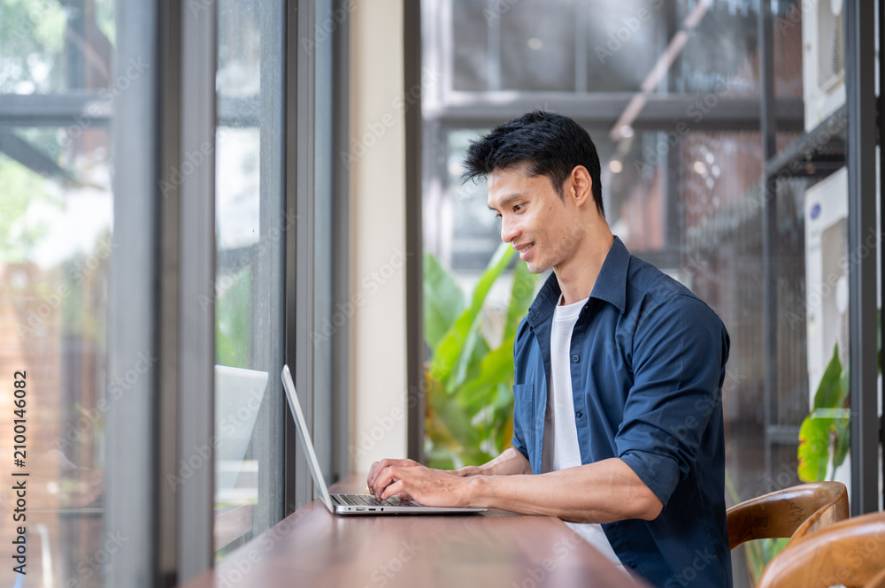 Fototapeta premium Young Asian freelancer working on laptop at wooden table beside glass wall in modern cafe