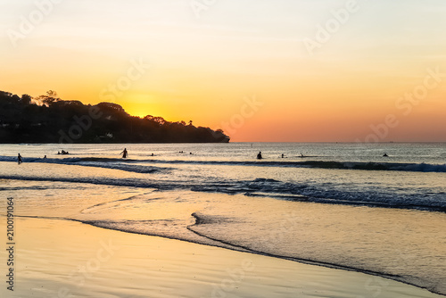 People Swimming in Ocean Waves at Tropical Beach During Vivid Orange Sunset with Tree Silhouette