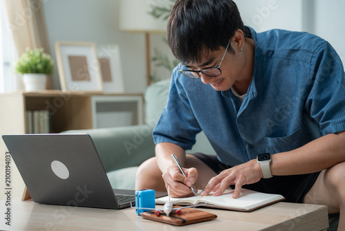 Man with glasses writing in notebook with laptop, miniature airplane and luggage, planning budget for next travel vacation at home