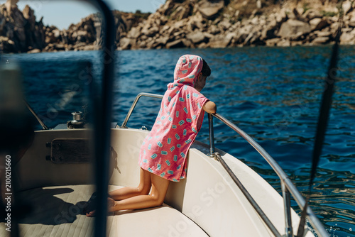 Child wrapped in a pink hooded towel poncho kneels at the bow of a boat facing a rocky coastline. Coastal boat excursions and family sea travel offering children adventure and discovery