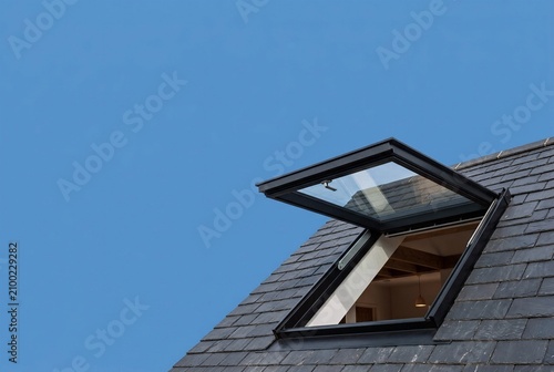 Open skylight window on a dark slate roof against a clear blue sky, revealing interior wooden beams and providing natural light.