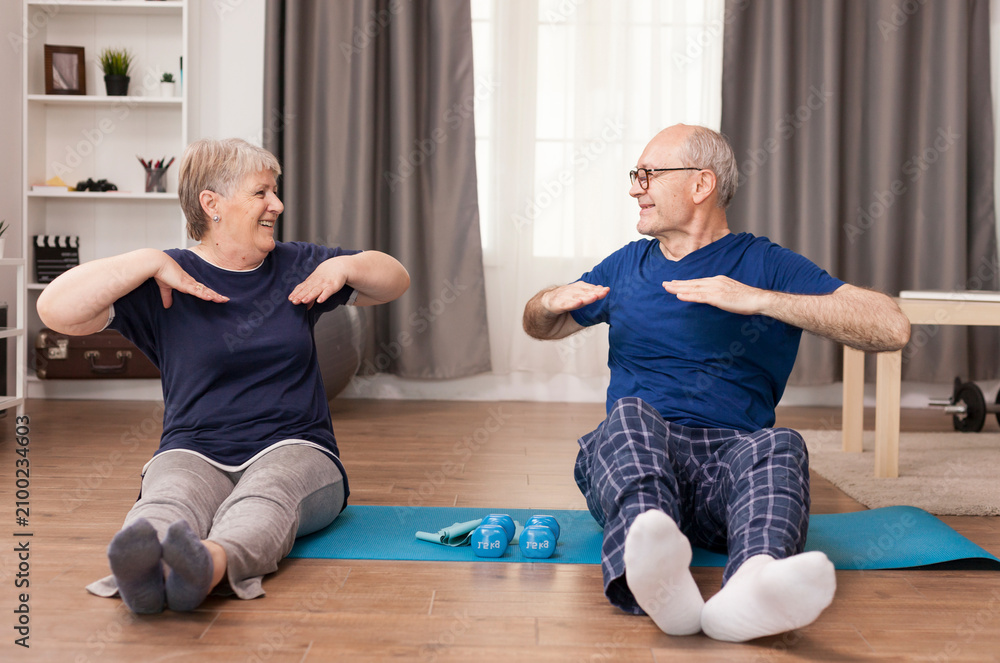Obraz premium Aged couple stretches on a yoga mat and smiles at each other, sharing a home workout and healthy retirement routine, combining couple exercise, healthy aging and shared home workout routine.