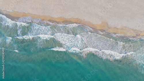 Horizontal aerial top down view of turquoise ocean waves washing on a sandy beach shore with sparkling white foam during a bright sunny summer day for travel promo