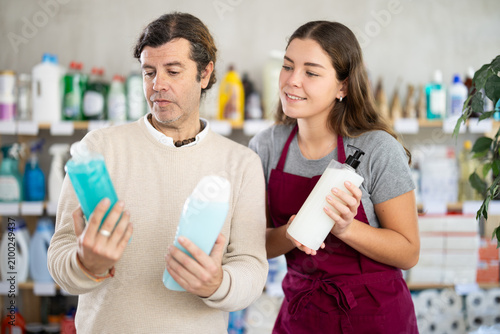 Smiling friendly young saleswoman offering various shampoo options to interested middle-aged man thoughtfully examining bottles in personal care store