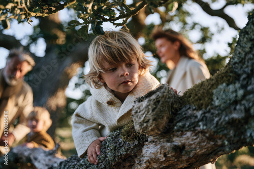 A young child curiously peers from a tree branch, surrounded by family members enjoying a serene moment in nature. The warmth of the sunlight enhances the peaceful scene.
