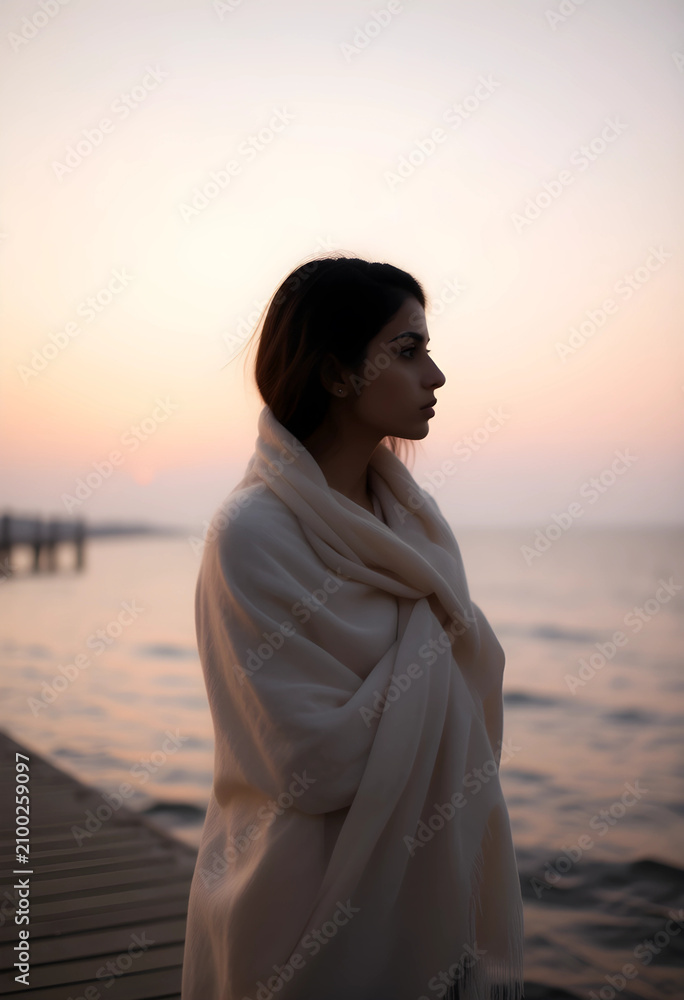 Fototapeta premium Young woman wrapped in a warm blanket standing on a wooden pier at sunset, looking out over the calm water