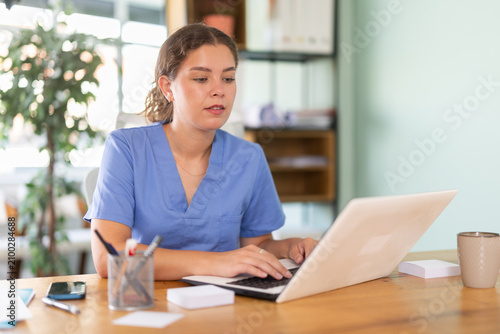 Quadro su tela Female physician in blue uniform filling up medical forms on laptop while sittin