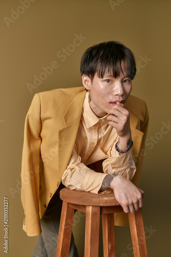 Bild auf Leinwand A stylish young Asian man leans on a stool, flaunting his chic outfit and confident vibe