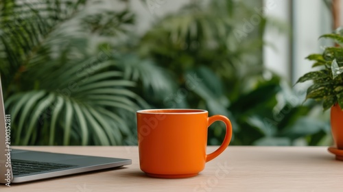 An orange coffee mug sits on a wooden desk next to a laptop, with lush green plants blurred in the background.
