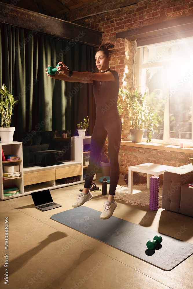 Obraz premium Athletic woman jumping with dumbbell during high intensity home workout. Concept of cardio strength training, calorie burning, online fitness and active lifestyle.