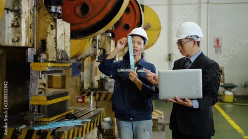 Asian female factory worker and male manager engineer in hard hats inspect a metal part near a stamping press machine. Quality control discussion with laptop in an industrial manufacturing plant.