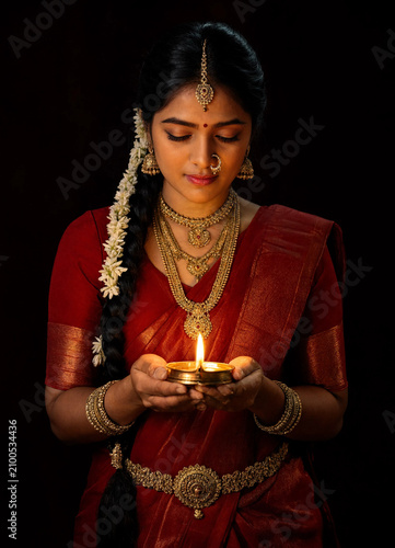 Portrait of a serene South Indian woman in a traditional red silk saree and gold jewelry holding a lit diya lamp, Diwali festival celebration