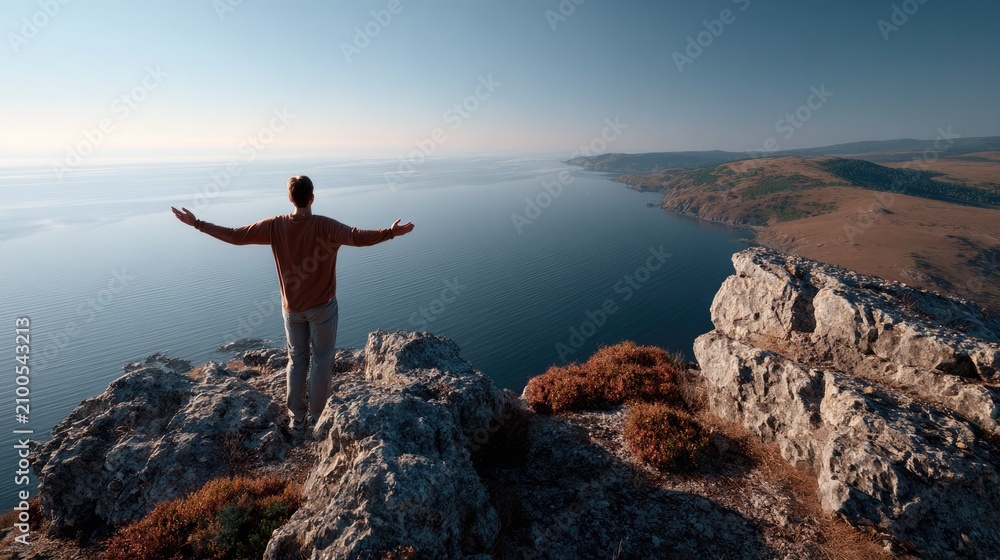 Obraz premium Serene Summit: A lone figure stands with arms outstretched on a rugged cliff edge, embracing the breathtaking panorama of the vast ocean and distant coastline under a clear, azure sky.