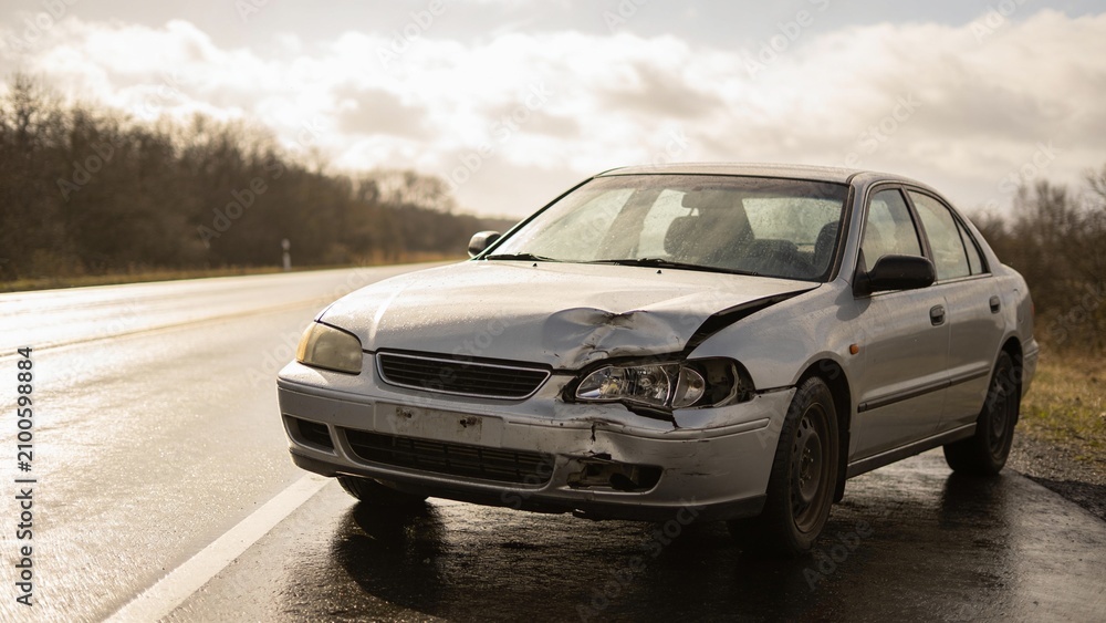 Fototapeta premium Damaged silver sedan abandoned on empty highway during dusk with trees in background