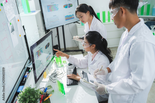 Laboratory scientist research computer analysis data teamwork experiment with Asian adult team in white coat studying chart, sample, glassware inside modern clinic lab