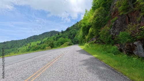 Driver's perspective along the Blue Ridge Parkway toward Waterrock Knob on a sunny day, surrounded by lush green forests in the North Carolina mountains