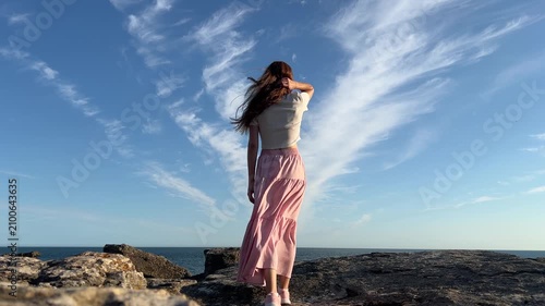 Woman in pink skirt walking toward an ocean cliff at sunset, reflection and travel lifestyle concept