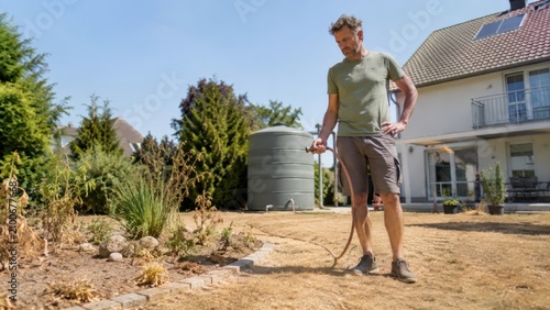 Mann steht mit Gartenschlauch vor vertrocknetem Rasen und dürren Pflanzen. Hitzewelle, Wassermangel und anhaltende Trockenheit verursachen schwere Schäden im heimischen Garten.