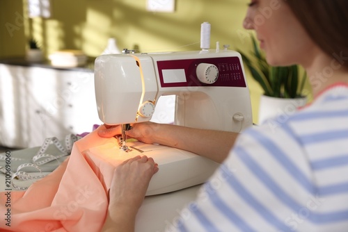 Woman sewing fabric with professional machine at white table in workshop, closeup