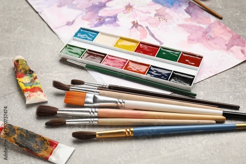 Фотография Different brushes, watercolor paints, tubes and drawing on grey table, closeup