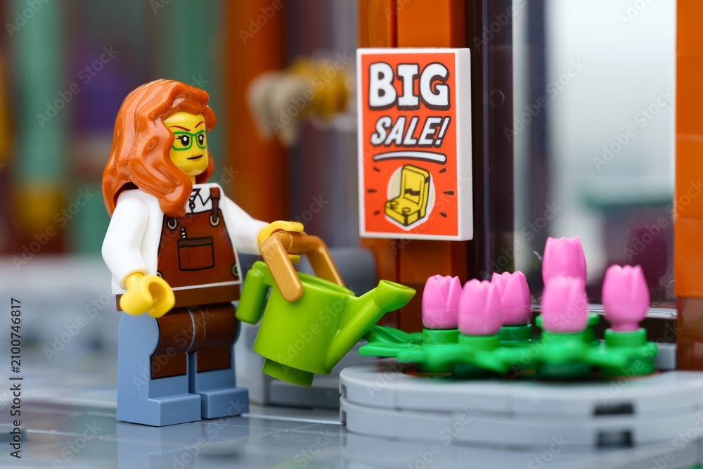 Fototapeta premium Tambov, Russian Federation - June 28, 2026 A Lego woman minifigure with red hair watering pink tulip flowers using a green watering can outside a store with a big sale sign.