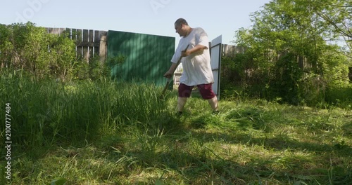 Caucasian man using a traditional hand scythe to mow overgrown tall grass in a rural backyard on a sunny summer day, engaging in agricultural work and lawn care with classic farming tool.